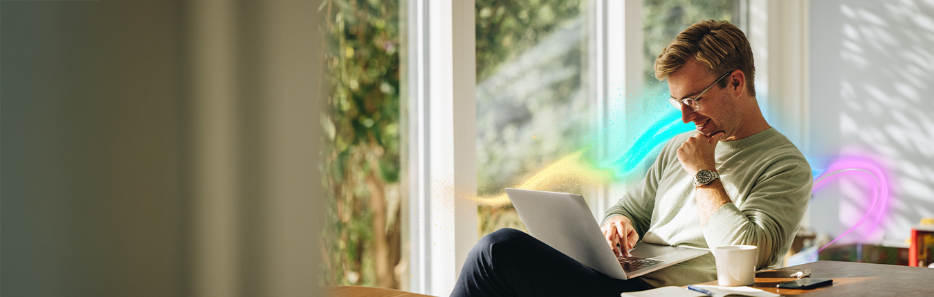 Uśmiechnięty mężczyzna siedzący przy stole, pracujący na laptopie. Na stole otwarty notatnik, kubek oraz smartfon.