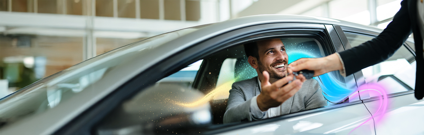 Uśmiechnięty mężczyzna, który wychyla się przez przednie okno samochodu i jednocześnie odbiera kluczyk od drugiej osoby. Sylwetki drugiej osoby nie widać.