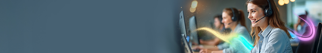 Uśmiechnięta kobieta rozmawia przez zestaw słuchawkowy. w biurze przed laptopem. W tle inne osoby rozmawiające przez zestawy słuchawkowe.