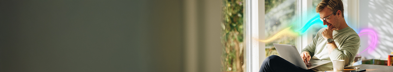 Uśmiechnięty mężczyzna siedzący przy stole, pracujący na laptopie. Na stole otwarty notatnik, kubek oraz smartfon.