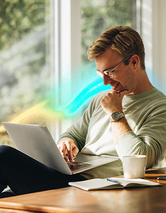 Uśmiechnięty mężczyzna siedzący przy stole, pracujący na laptopie. Na stole otwarty notatnik, kubek oraz smartfon.