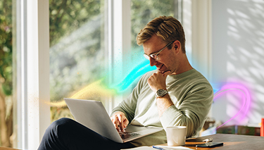 Uśmiechnięty mężczyzna siedzący przy stole, pracujący na laptopie. Na stole otwarty notatnik, kubek oraz smartfon.