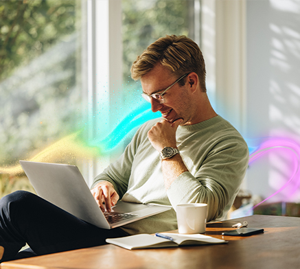 Uśmiechnięty mężczyzna siedzący przy stole, pracujący na laptopie. Na stole otwarty notatnik, kubek oraz smartfon.