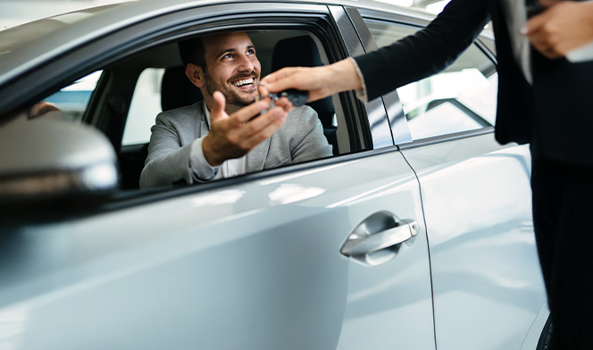 Uśmiechnięty mężczyzna, który wychyla się przez przednie okno samochodu i jednocześnie odbiera kluczyk od drugiej osoby. Sylwetki drugiej osoby nie widać.
