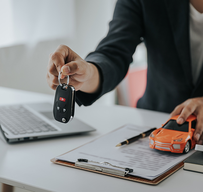 Dłoń mężczyzny przekazująca kluczyki do samochodu. W tle biurko z laptopem, dokumentami oraz modelem samochodu.