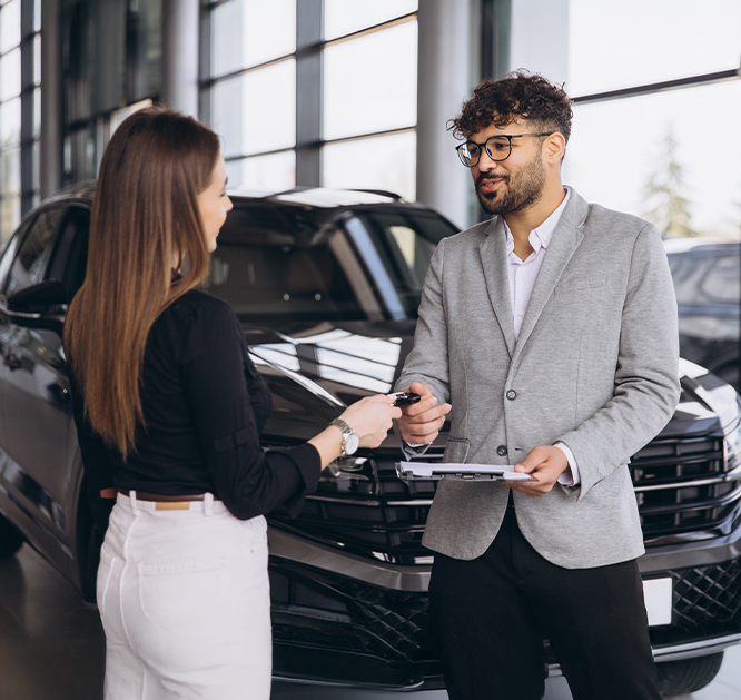 Uśmiechnięty mężczyzna przekazujący kluczyki do samochodu kobiecie. Znajdują się w salonie samochodowym. W tle samochód.
