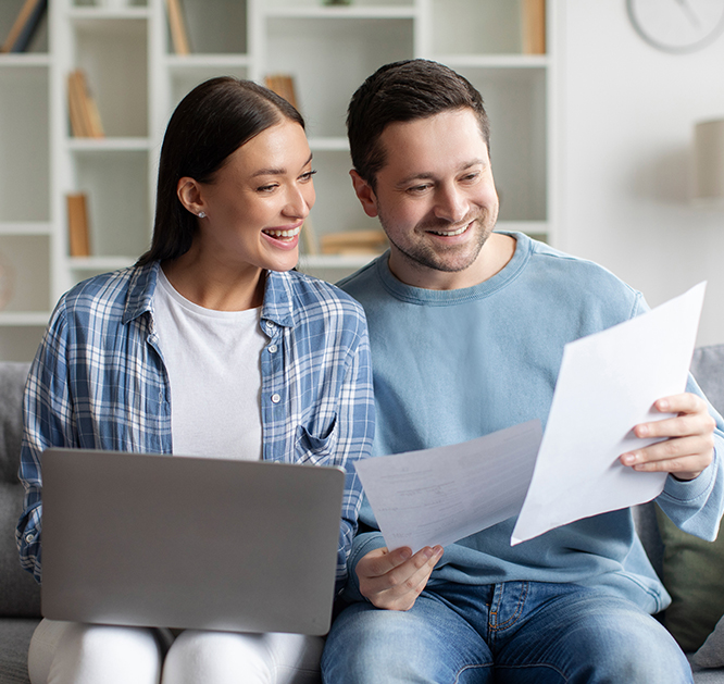 Uśmiechnięta kobieta i uśmiechnięty mężczyzna siedzą na kanapie i czytają dokumenty. Kobieta ma laptopa na kolanach.