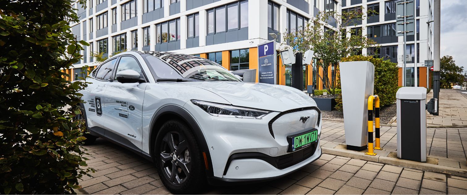 Biały Ford Mustan wersja elektryczna, wyjeżdża z parkingu w tle biurowce.