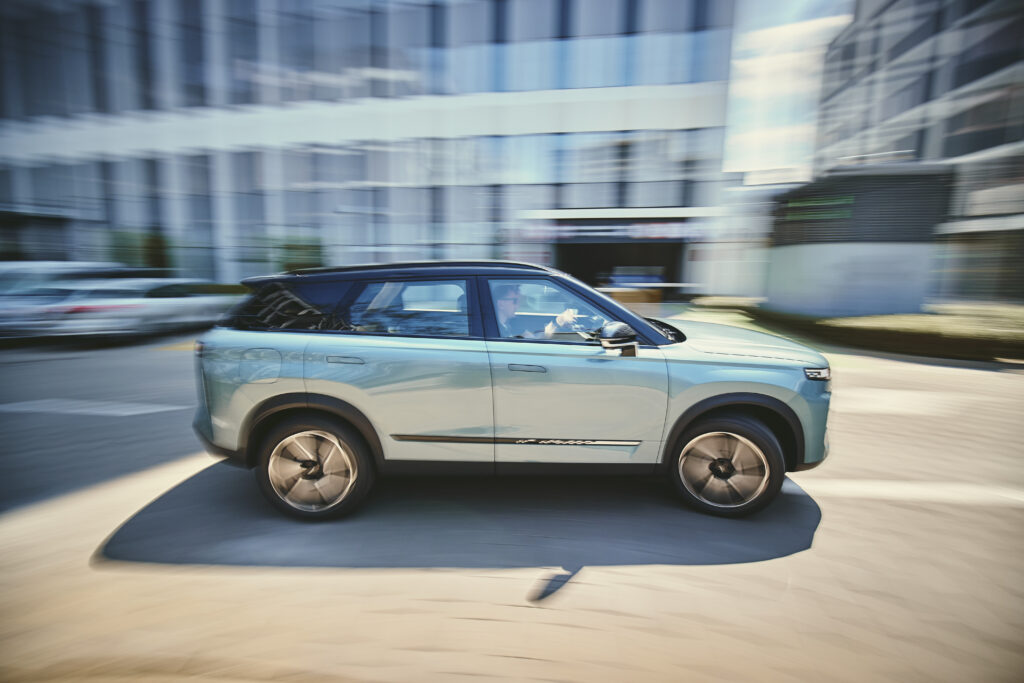 Srebrne auto marki Jaecoo jedzie ulicą, w tle biurowce.