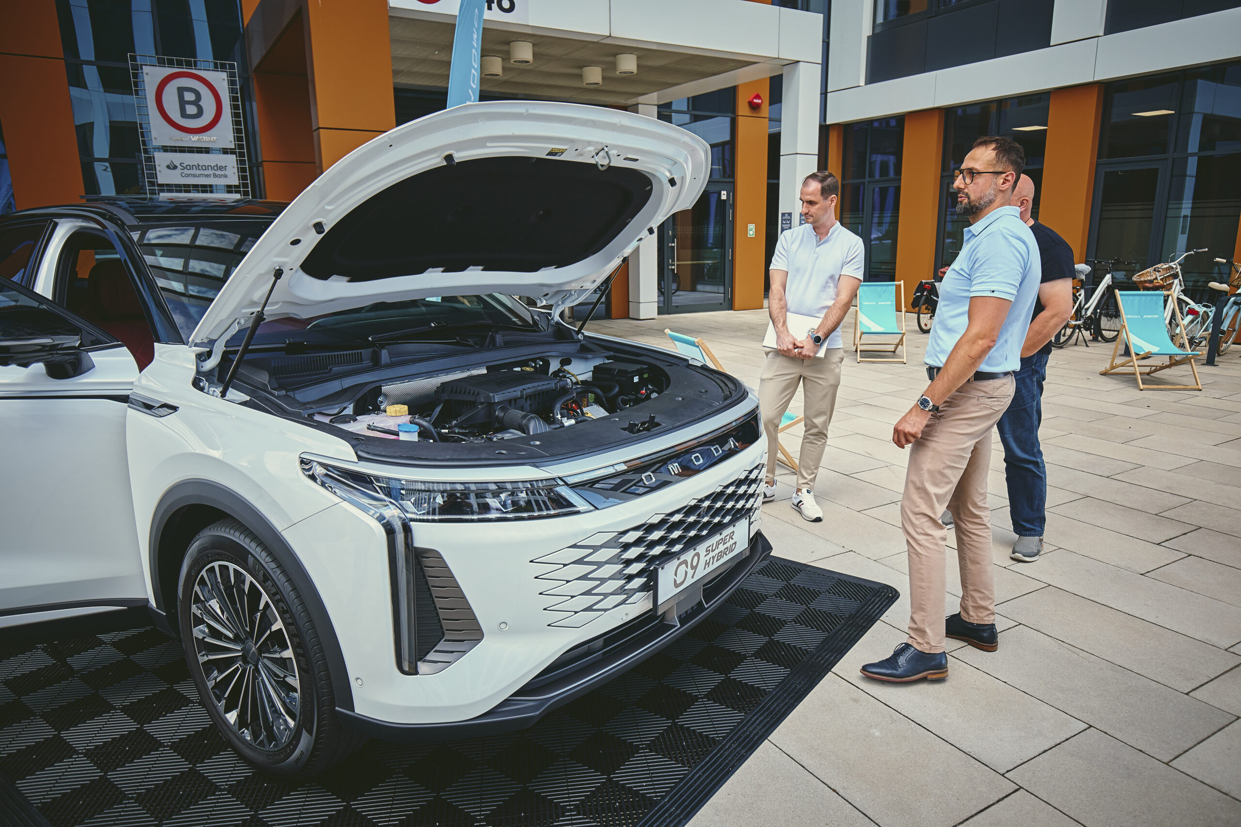 Na pierweszym planie białe auto marki OMODA z otwartą klapą, przy nim trzech mężczyzn przyglądających się mu