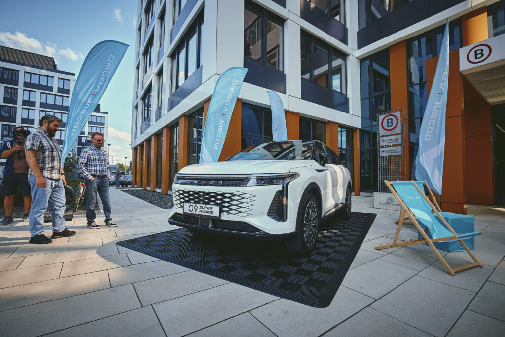 Białe auto marki omoda stoi na parkingu, wokół auta flagi z logotypem OMODA i osoby przyglądające się mu