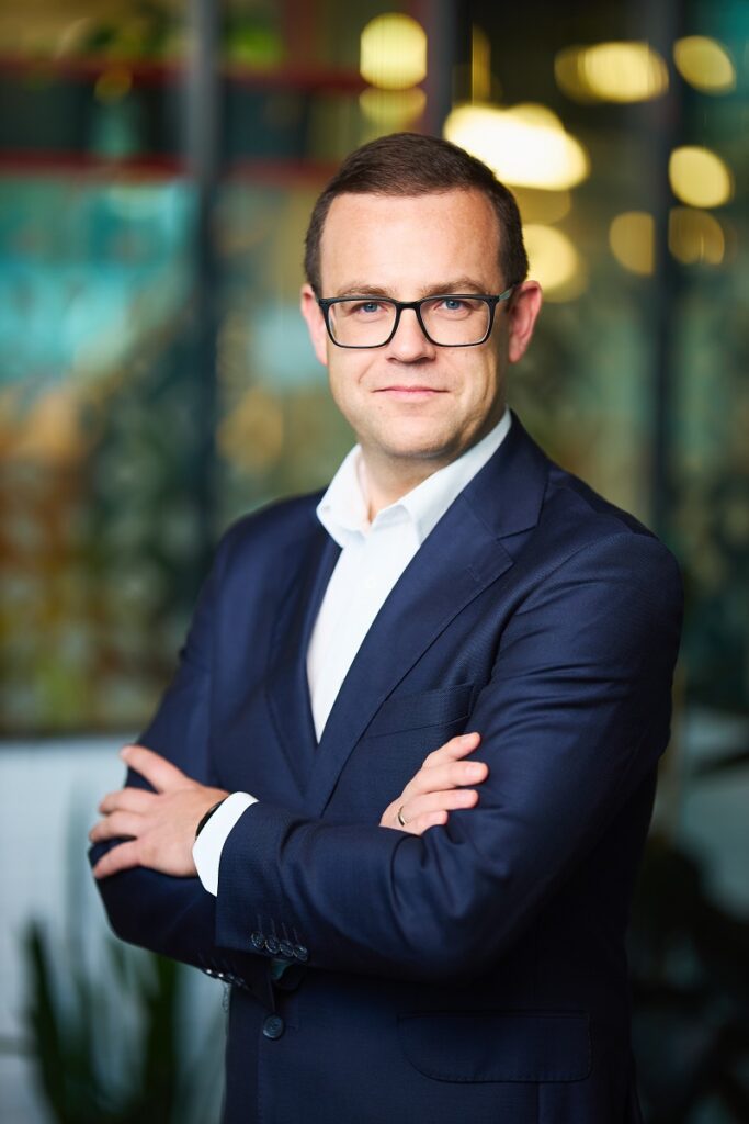 Młodszy mężczyzna w okularach patrzący w kamerę z lekkim uśmiechem