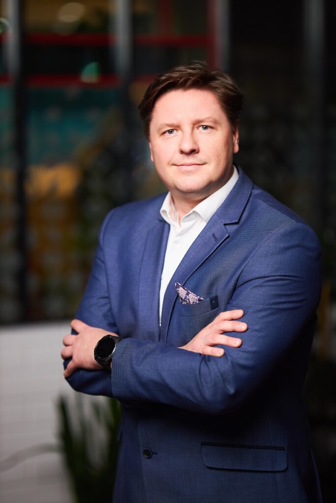 Młodszy mężczyzna w garniturze patrzący w kamerę z założonymi rękami