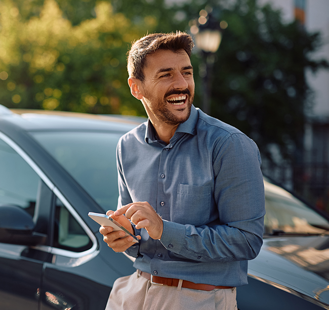 Uśmiechnięty mężczyzna idący obok samochodu. Trzyma w dłoniach smartfona.