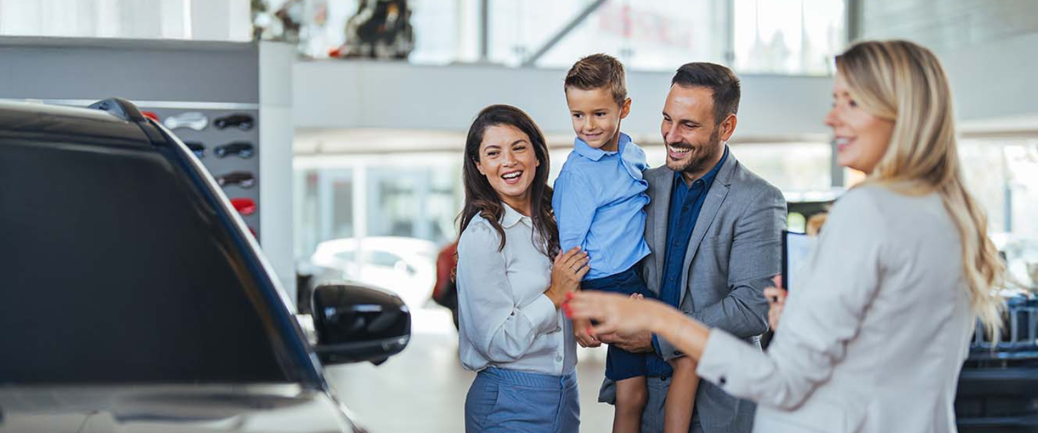 Uśmiechnięta rodzina trzyosobowa w salonie samochodowym ogląda auta z doradcą